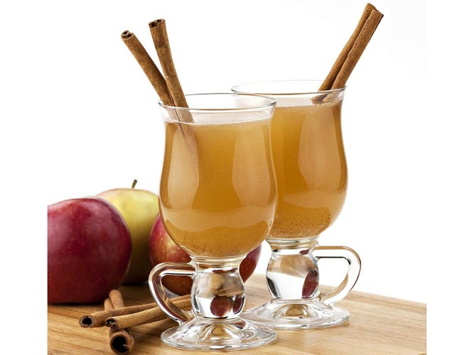 Two glasses of apple cider with cinnamon sticks, with apples and cinnamon powder in the background.