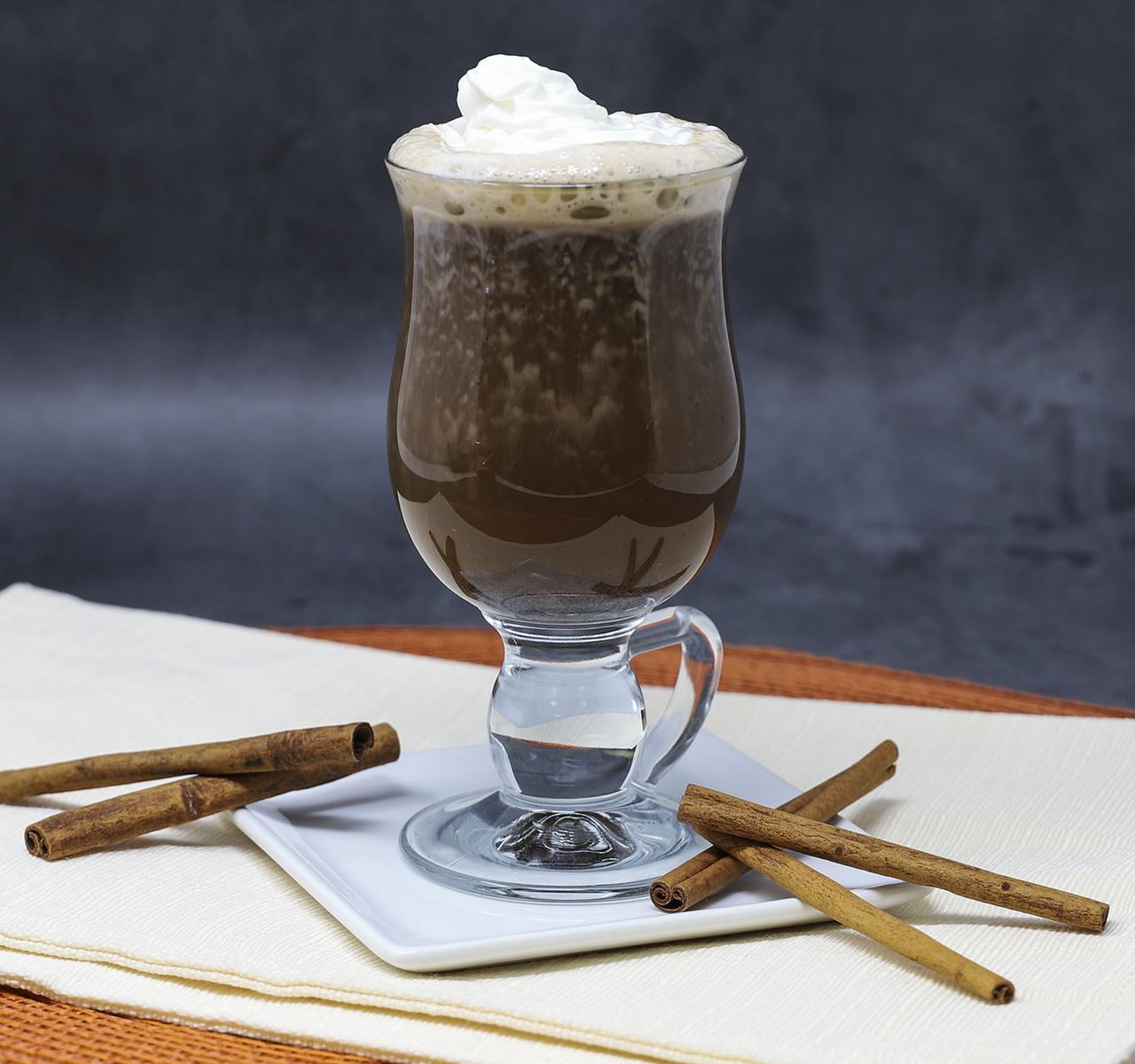 Chai tea with whipped cream in a glass, surrounded by cinnamon sticks on a dark background