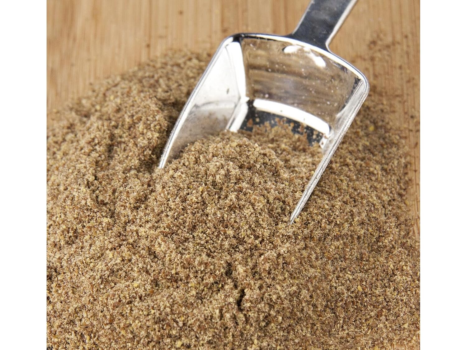 A close-up of a pile of fine brown brown flaxseed meal with a metal scoop resting in the center on a light wood surface.